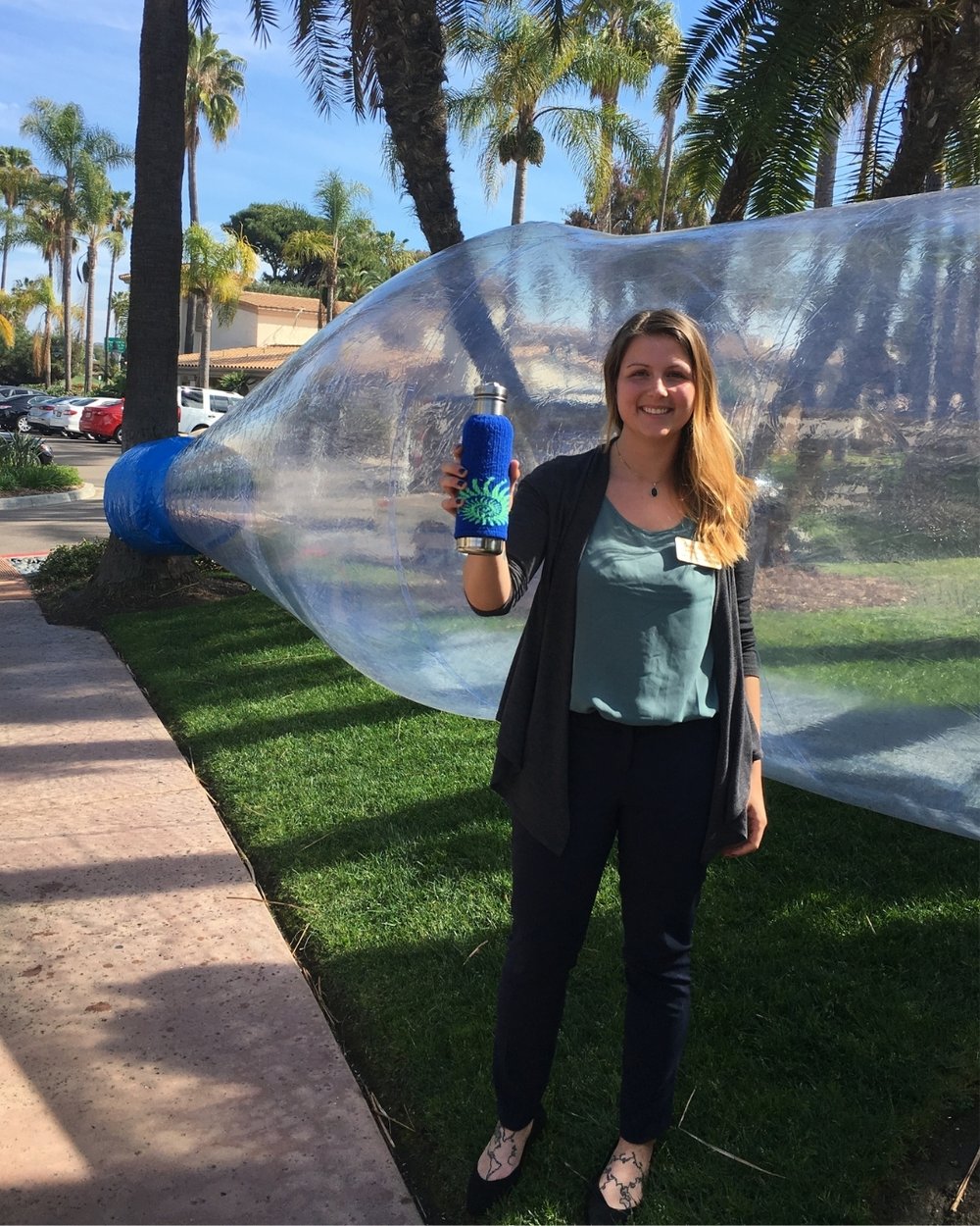 Sam poses in front of a plastic water bottle the size of a whale. She is holding a reusable water bottle in her hand.