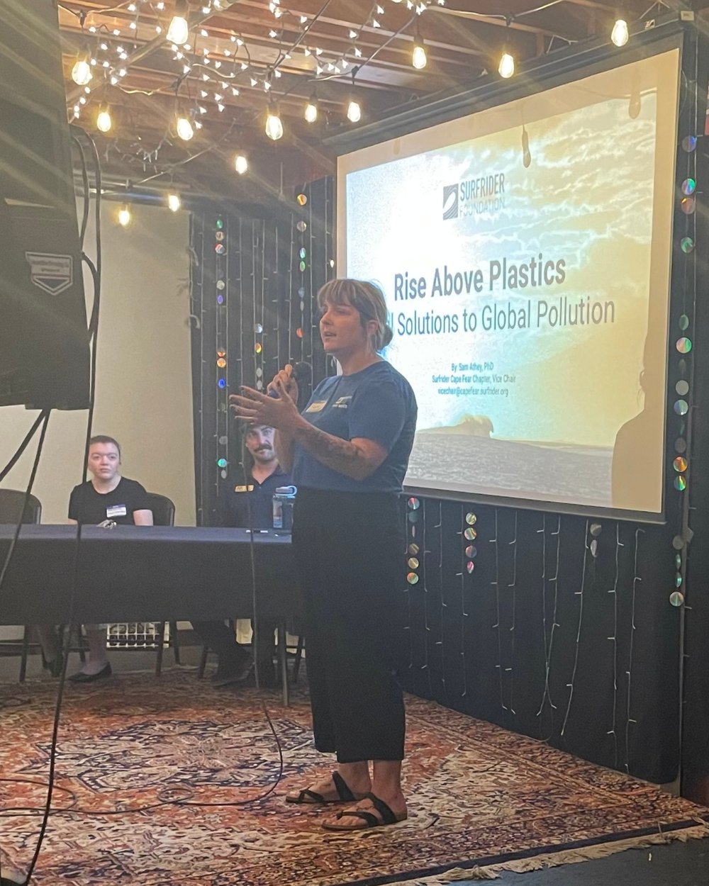 Sam presents in front of an audience, standing on a colorful rug. The powerpoint reads, 'Rise Above Plastics.'
