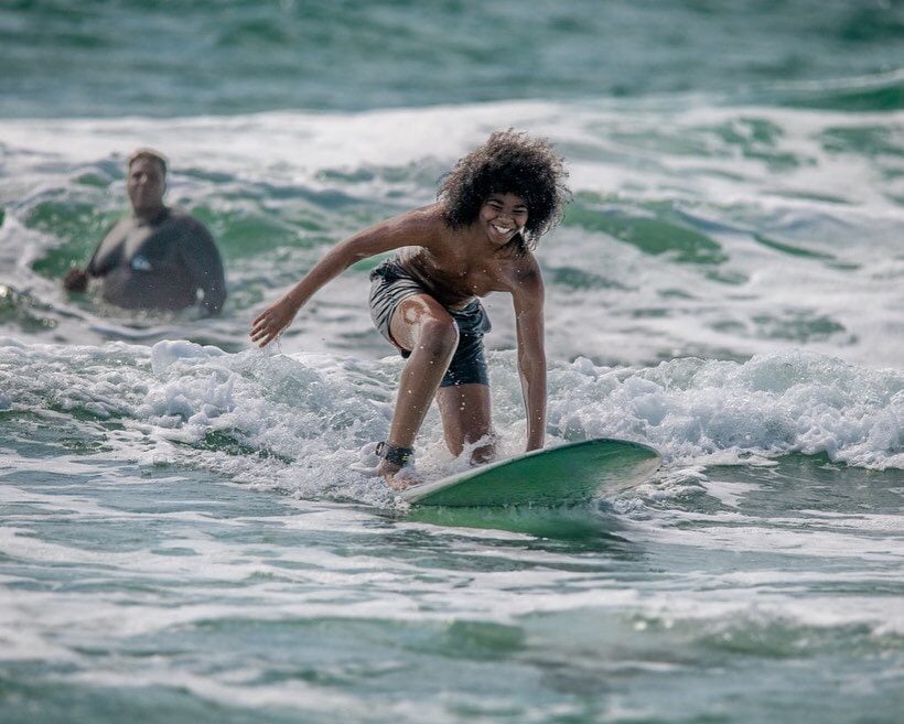 A smiling Black youth works to stand up on a surfboard after being pushed into a wave at Wrightsville Beach.